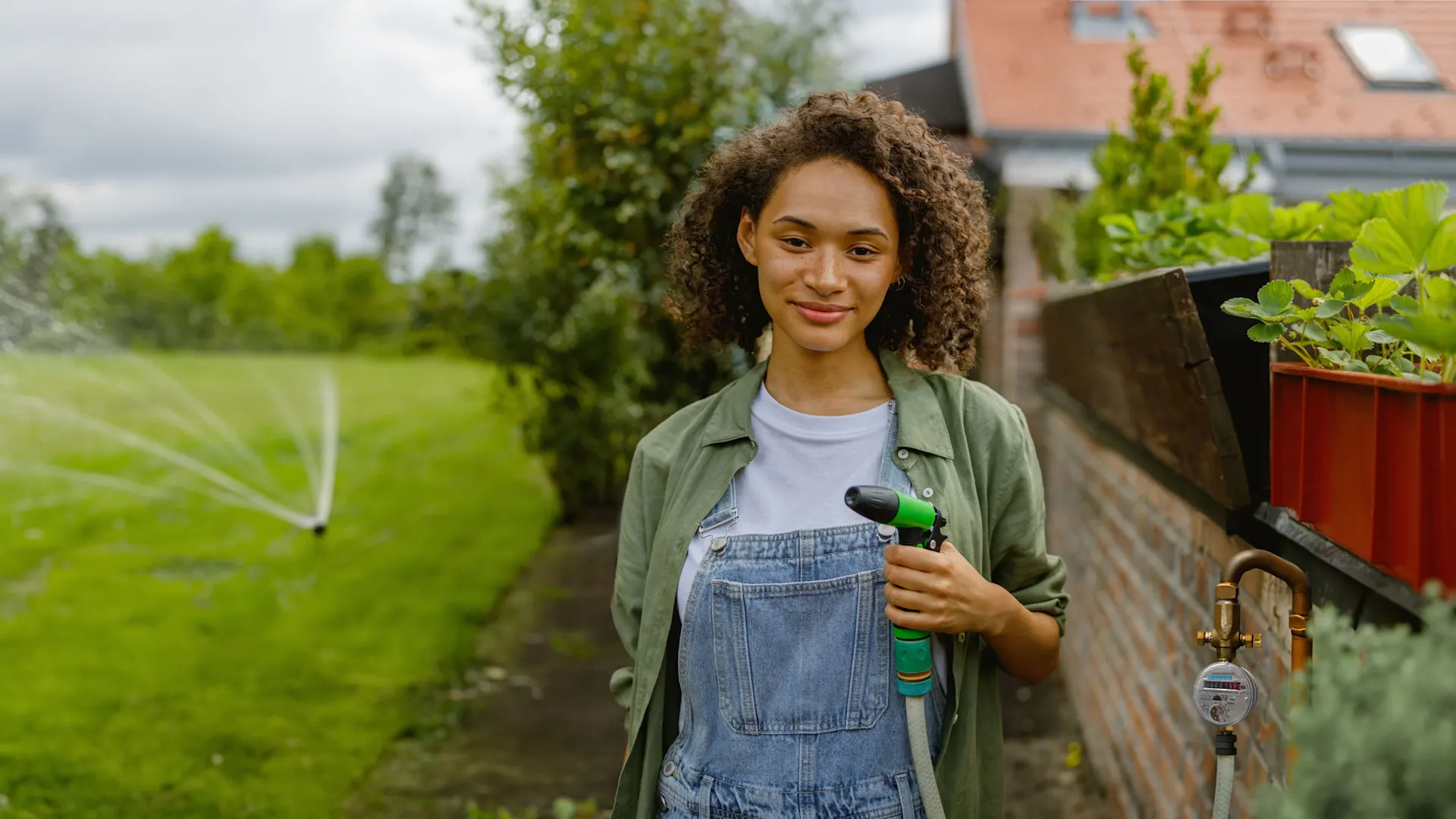 Gartenwasserzähler Eine Frau steht an einer Mauer im Garten, mit einem Gartenschlauch in der linken Hand. An der Mauer ist ein Gartenwasserzähler der NEW. Auf der Wiese ist ein Wassersprenger.
