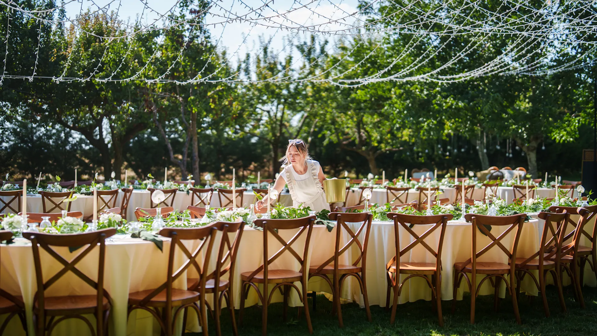 NEW Magazin Hochzeitslocations Zwei große lange Tischreihen stehen in einem sonnigen Garten mit Bäumen. Eine Frau deckt ein. Die Tische sind festlich geschmückt, Lichterketten hängen darüber. Credit: Ugur Karakoc