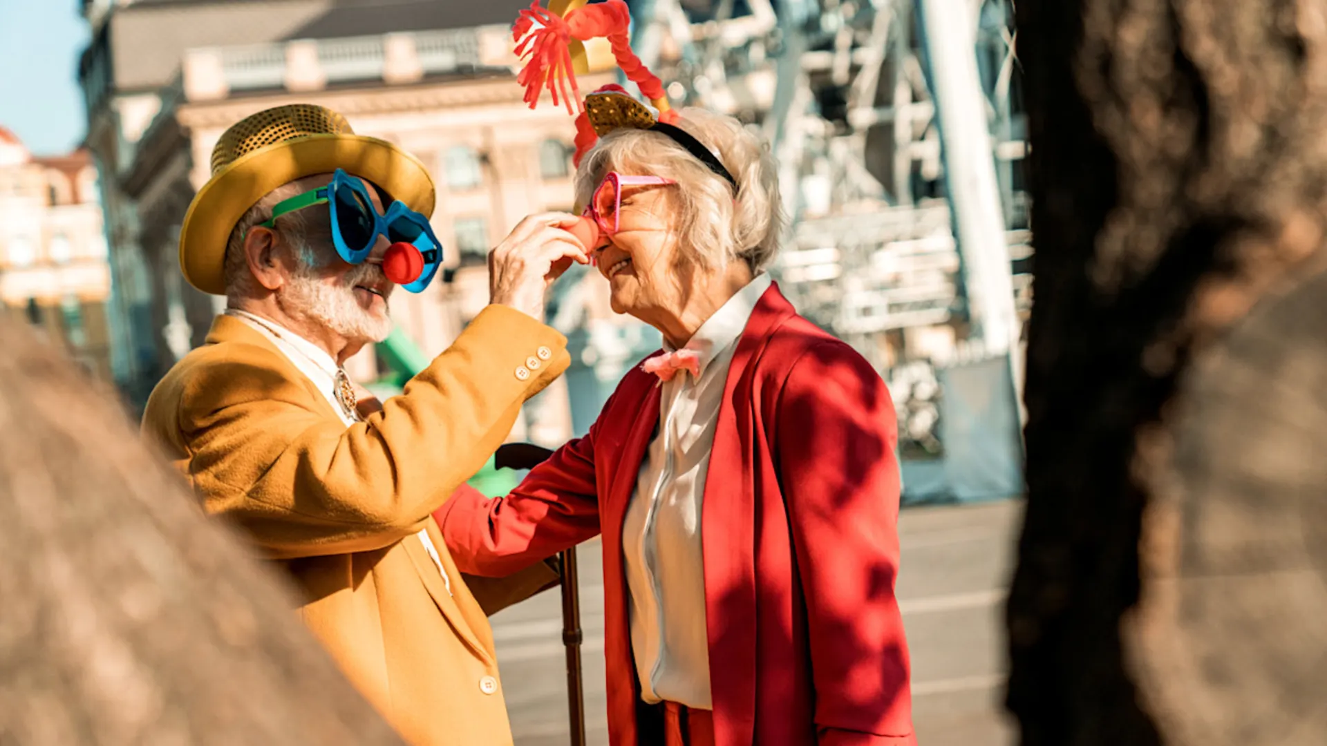 Karnevalsfeiern weltweit Das Bild zeigt ein älteres Pärchen. Der Mann links trägt einen goldenen Hut, eine senfgelbe Jacke und eine blaue Clownbrille. Die Frau rechts trägt einen roten Blazer, ein weißes Hemd und eine rote Kopfdekoration. Beide lachen sich an.