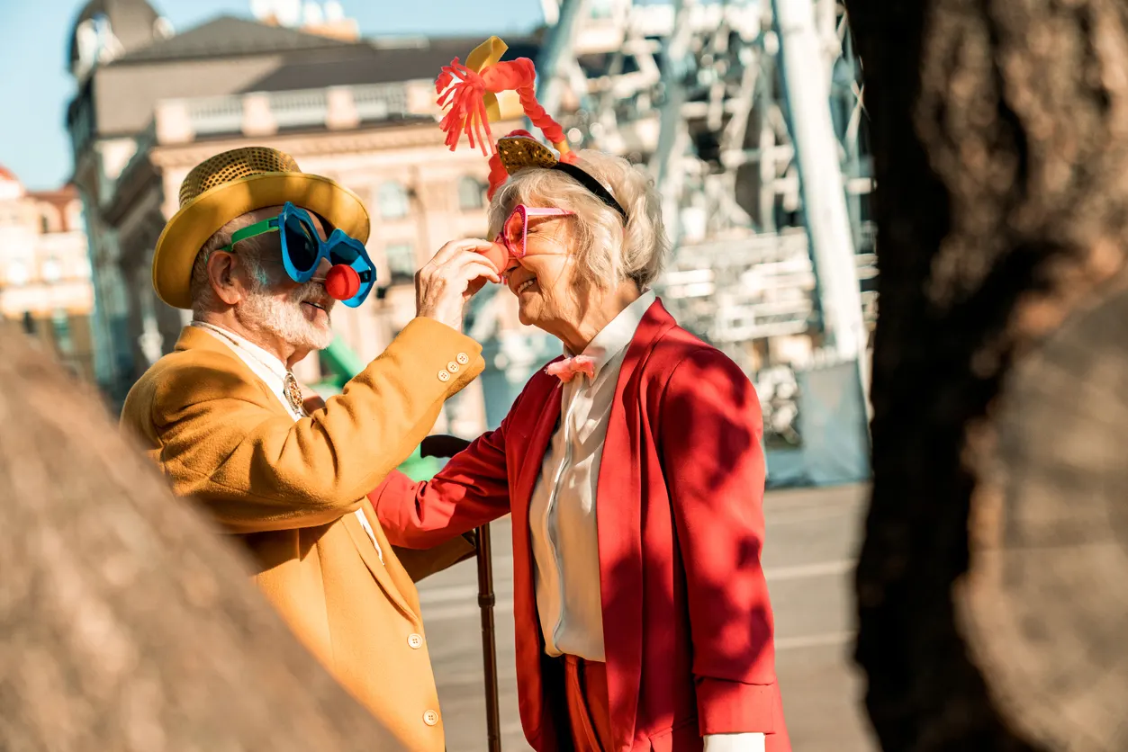 Das Bild zeigt ein älteres Pärchen. Der Mann links trägt einen goldenen Hut, eine senfgelbe Jacke und eine blaue Clownbrille. Die Frau rechts trägt einen roten Blazer, ein weißes Hemd und eine rote Kopfdekoration. Beide lachen sich an.