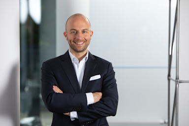 Ein Mann mit Glatze und Bart in dunklem Anzug mit weißem Hemd und Einstecktuch steht lächelnd mit verschränkten Armen in einem modernen Büro. Im Hintergrund sind helle Glaswände und verschwommene Pflanzen zu sehen. Ein Mann mit Glatze und Bart in dunklem Anzug mit weißem Hemd und Einstecktuch steht lächelnd mit verschränkten Armen in einem modernen Büro. Im Hintergrund sind helle Glaswände und verschwommene Pflanzen zu sehen.