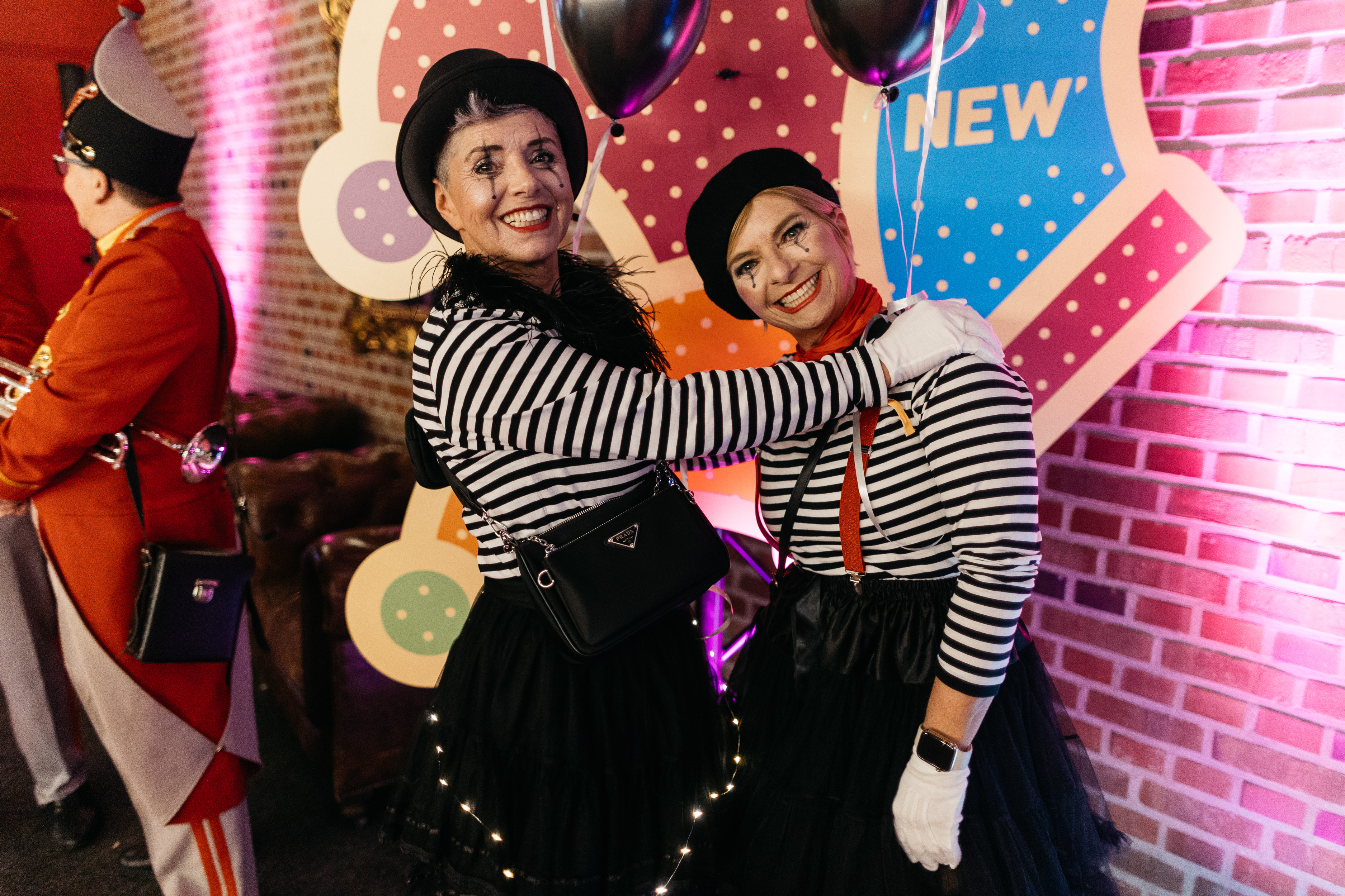 Zwei Personen in schwarz-weiß gestreiften Oberteilen und schwarzen Hosen umarmen sich vor einer bunten Kulisse mit gepunktetem Hintergrund. Beide tragen schwarze Hüte und weiße Handschuhe.