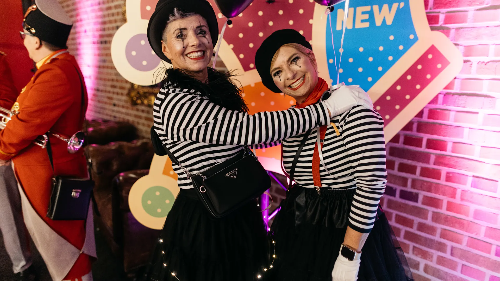 Zwei Personen in schwarz-weiß gestreiften Oberteilen und schwarzen Hosen umarmen sich vor einer bunten Kulisse mit gepunktetem Hintergrund. Beide tragen schwarze Hüte und weiße Handschuhe. Zwei Personen in schwarz-weiß gestreiften Oberteilen und schwarzen Hosen umarmen sich vor einer bunten Kulisse mit gepunktetem Hintergrund. Beide tragen schwarze Hüte und weiße Handschuhe.