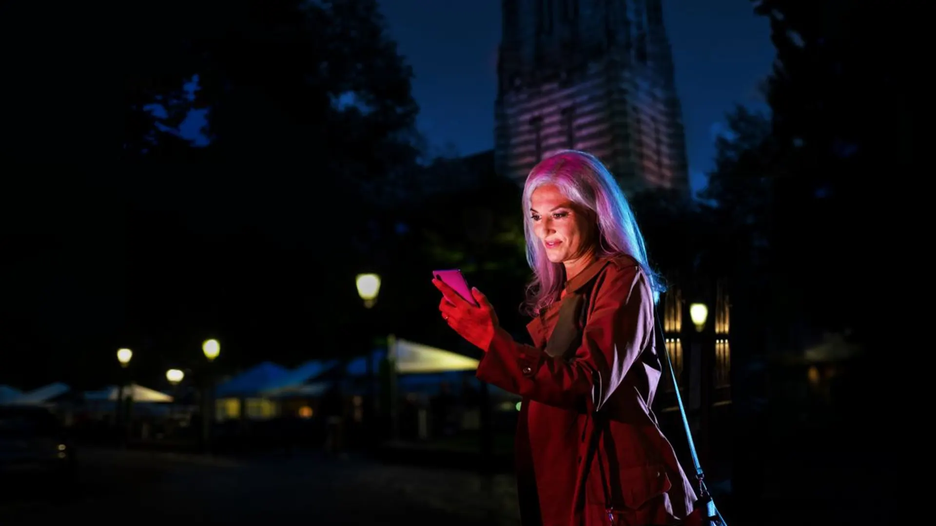 Nachts unterwegs mit der NEW Das Bild zeigt eine junge Frau, die im Dunkeln auf einer Straße unterwegs ist. Sie trägt eine braune Jacke und hält ein Mobiltelefon in der Hand. Im Hintergrund sind Häuser, Laternen und Dekorationen zu sehen.
