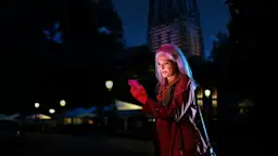 Nachts unterwegs mit der NEW Das Bild zeigt eine junge Frau, die im Dunkeln auf einer Straße unterwegs ist. Sie trägt eine braune Jacke und hält ein Mobiltelefon in der Hand. Im Hintergrund sind Häuser, Laternen und Dekorationen zu sehen.