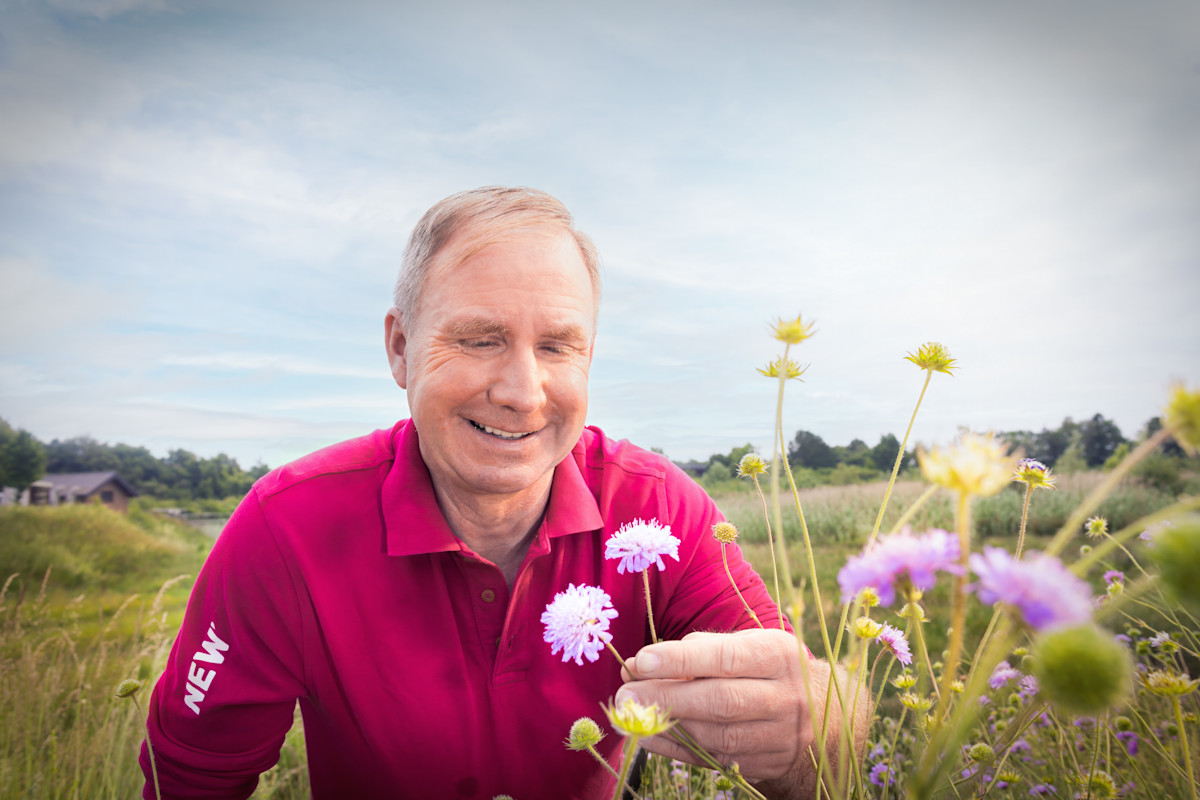 Ein lächelnder Mann in pinkem Poloshirt hält weiße Blumen in der Hand und steht in einer bunten Blumenwiese. Der Himmel ist bewölkt. Im Hintergrund sind grüne Felder und Bäume zu sehen.