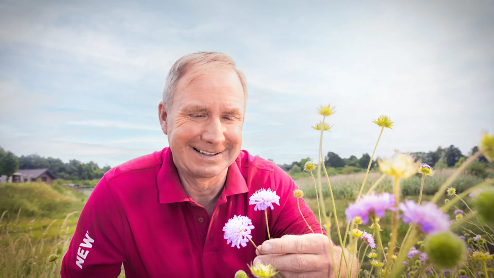 Ein Mitarbeiter auf einer Wildwiese in einem Wasserschutzgebiet Ein lächelnder Mann in pinkem Poloshirt hält weiße Blumen in der Hand und steht in einer bunten Blumenwiese. Der Himmel ist bewölkt. Im Hintergrund sind grüne Felder und Bäume zu sehen.