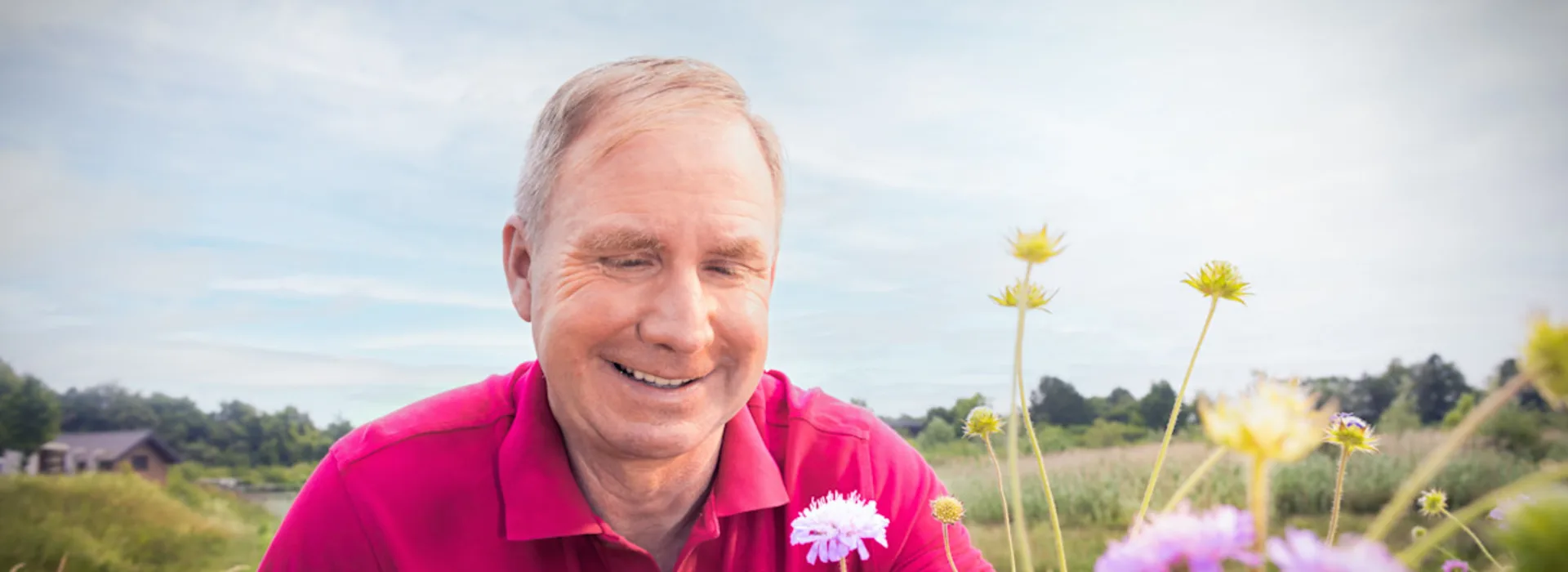 Ein Mitarbeiter auf einer Wildwiese in einem Wasserschutzgebiet Ein lächelnder Mann in pinkem Poloshirt hält weiße Blumen in der Hand und steht in einer bunten Blumenwiese. Der Himmel ist bewölkt. Im Hintergrund sind grüne Felder und Bäume zu sehen.