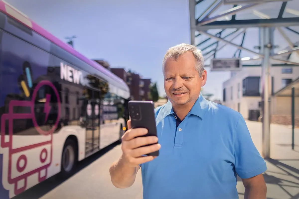 Ein Mann steht an einer Bushaltestelle und schaut in sein Smartphone. Ein NEW Bus steht im Hintergrund.