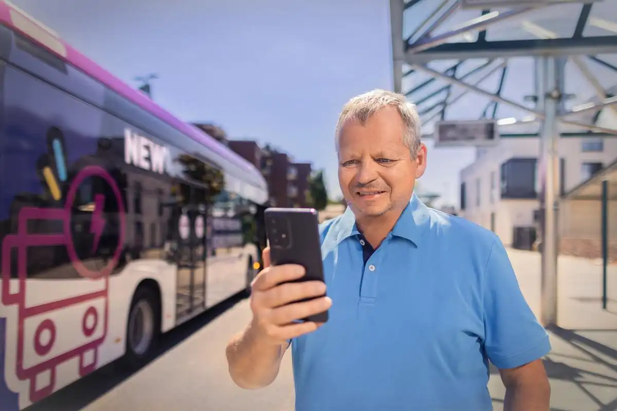 Ein Mann steht an einer Bushaltestelle und schaut in sein Smartphone. Ein NEW Bus steht im Hintergrund.