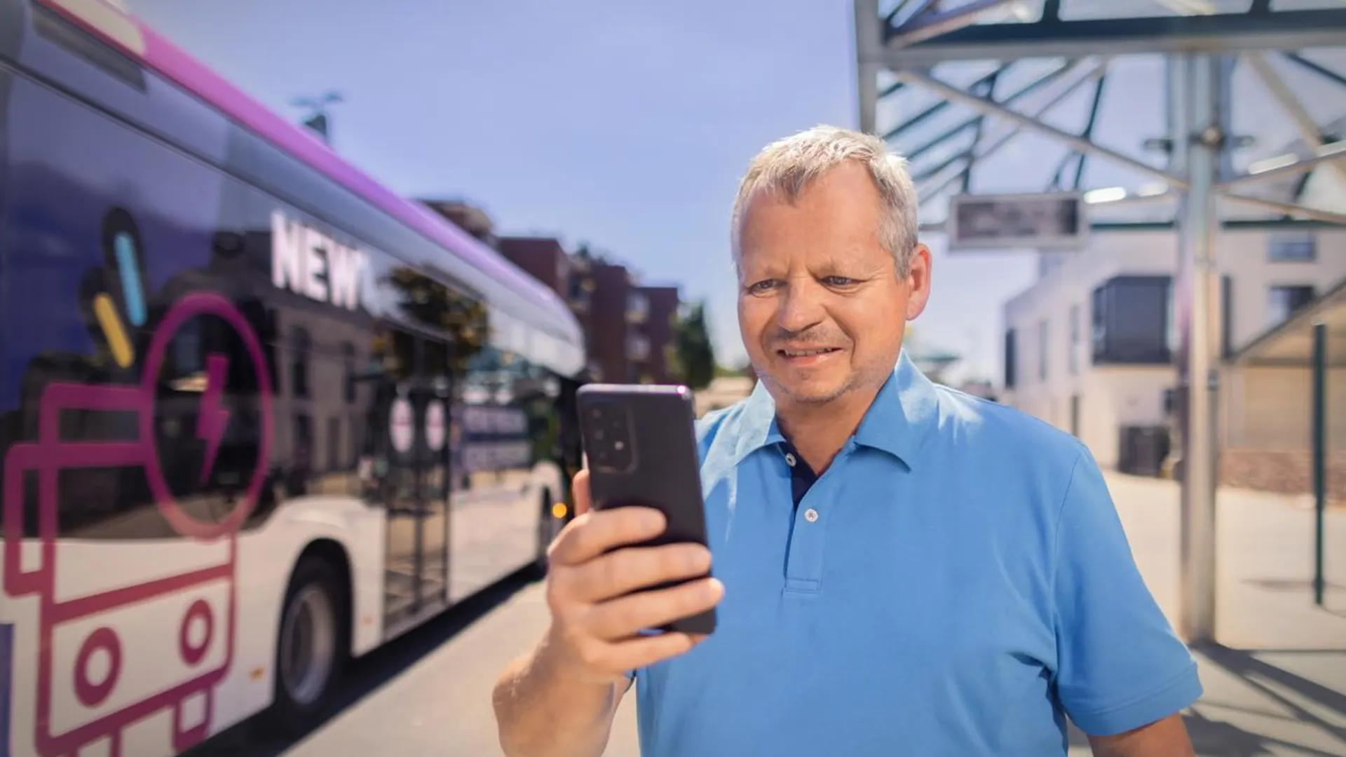 NEW MöBus Ein Mann steht an einer Bushaltestelle und schaut in sein Smartphone. Ein NEW Bus steht im Hintergrund.