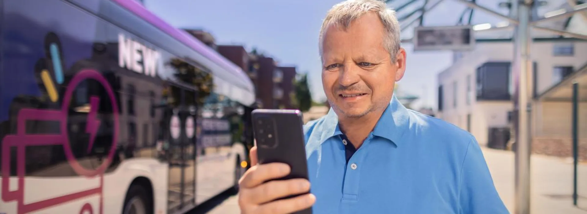 NEW MöBus Ein Mann steht an einer Bushaltestelle und schaut in sein Smartphone. Ein NEW Bus steht im Hintergrund.