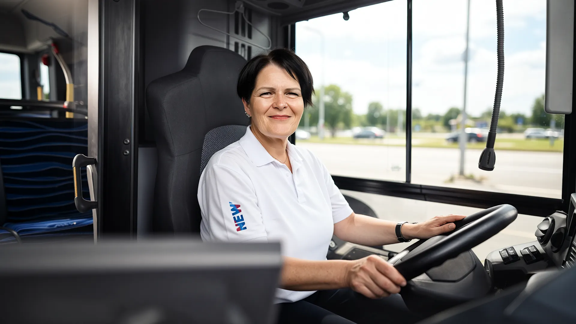 Manuela am Steuer eines Linienbusses Eine freundlich lächelnde Busfahrerin in weißem Poloshirt sitzt am Steuer eines modernen Busses. Durch die großen Fenster neben ihr ist eine sonnige Straßenlandschaft mit grünen Bäumen zu sehen. Die Fahrerin trägt das NEW-Logo auf ihrer Arbeitskleidung.