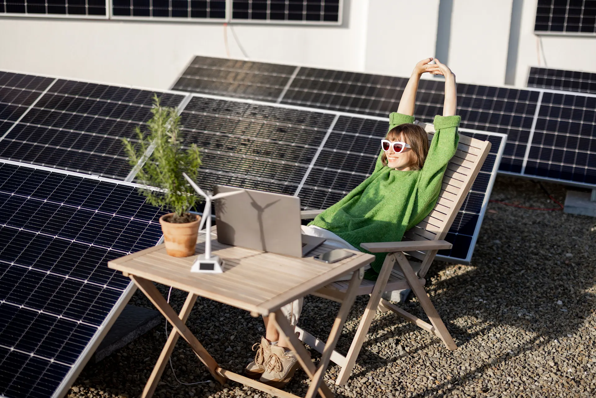 Eine Frau mit grünem Pullover und weißer Sonnenbrille entspannt sich auf einem Holzstuhl neben einem Tisch, auf dem sich ein Laptop, ein Miniatur-Windrad und eine Pflanze befinden. Sie ist von Solarpanelen umgeben. 