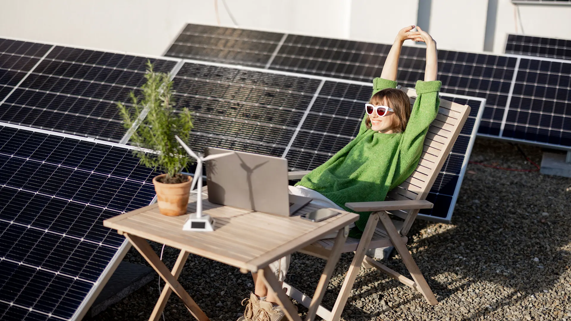 Strom selber erzeugen zu Hause Eine Frau mit grünem Pullover und weißer Sonnenbrille entspannt sich auf einem Holzstuhl neben einem Tisch, auf dem sich ein Laptop, ein Miniatur-Windrad und eine Pflanze befinden. Sie ist von Solarpanelen umgeben.