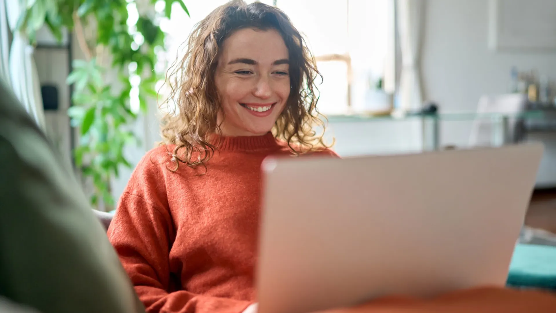 Studentin sitzt vor einem Laptop Eine junge Frau sitzt entspannt auf einem Sofa und lächelt, während sie auf ihren Laptop schaut. Im Hintergrund ist eine helle, wohnliche Umgebung mit Pflanzen zu sehen.