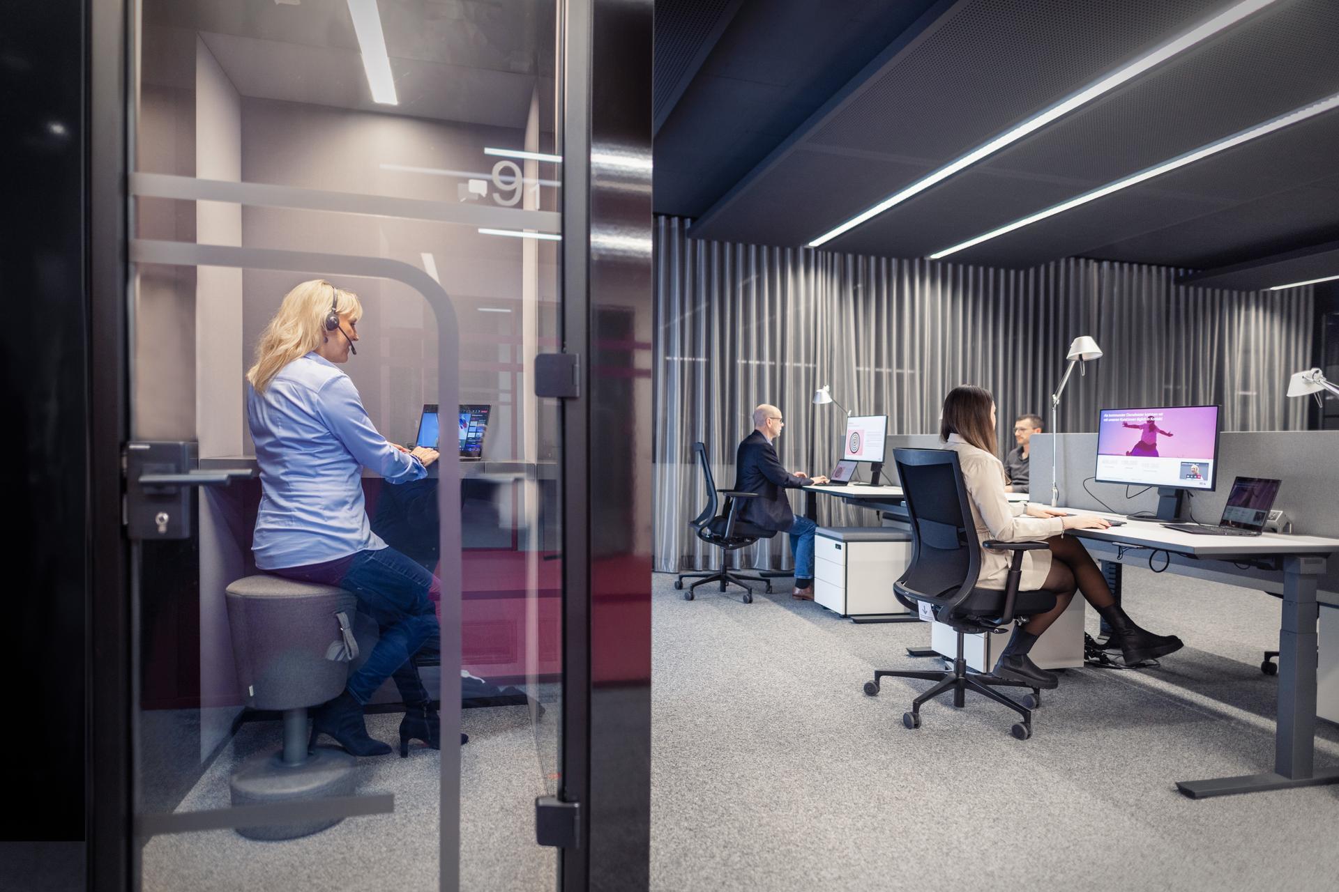 Modernes Büro mit einer Mitarbeiterin in einer gläsernen Telefonkabine und zwei Kolleg:innen, die an Schreibtischen im Hintergrund arbeiten. Die Räumlichkeiten sind in gedämpften Grau- und Blautönen gestaltet mit linearer LED-Beleuchtung an der Decke.
