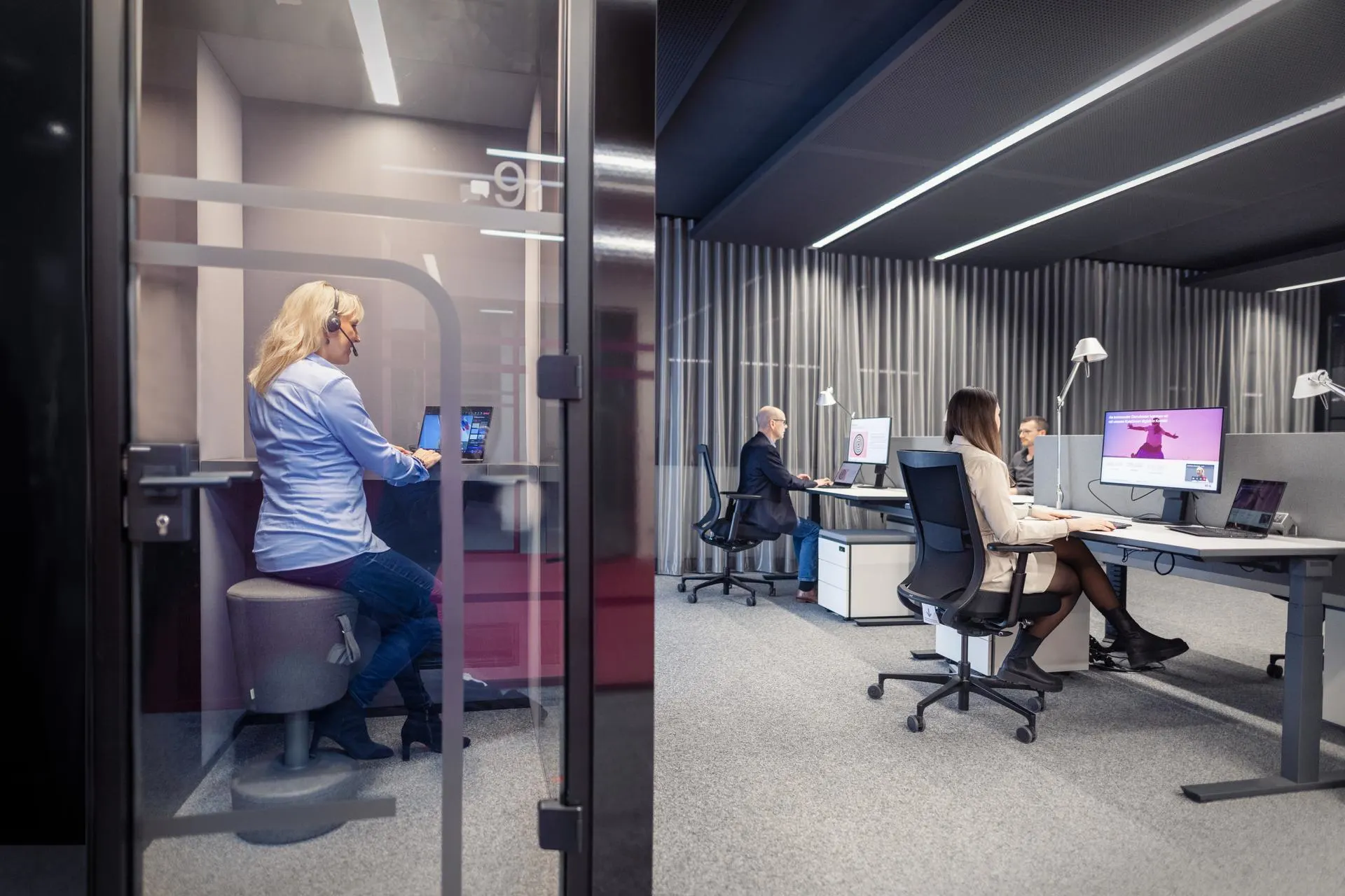 Modernes Büro mit einer Mitarbeiterin in einer gläsernen Telefonkabine und zwei Kolleg:innen, die an Schreibtischen im Hintergrund arbeiten. Die Räumlichkeiten sind in gedämpften Grau- und Blautönen gestaltet mit linearer LED-Beleuchtung an der Decke.