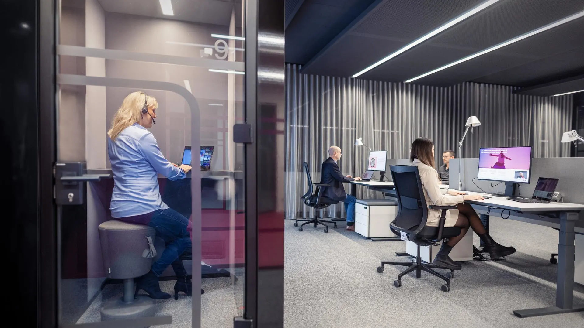 Papierloses Büro Modernes Büro mit einer Mitarbeiterin in einer gläsernen Telefonkabine und zwei Kolleg:innen, die an Schreibtischen im Hintergrund arbeiten. Die Räumlichkeiten sind in gedämpften Grau- und Blautönen gestaltet mit linearer LED-Beleuchtung an der Decke.
