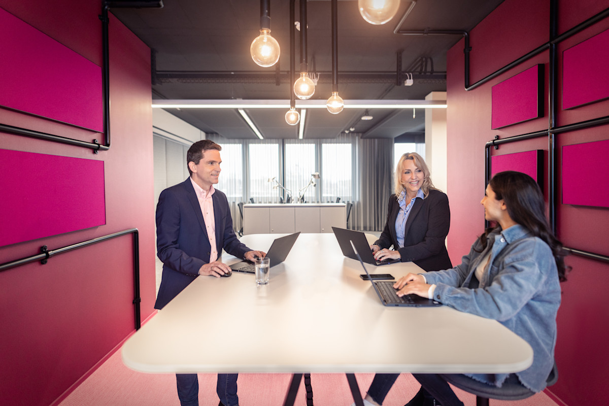 Drei Personen sitzen in einem modernen Besprechungsraum an einem weißen Tisch mit Laptops und führen ein Gespräch. Der Raum ist in auffälligem Pink gestaltet mit auffälligen Leuchtmitteln und großen Fenstern im Hintergrund.