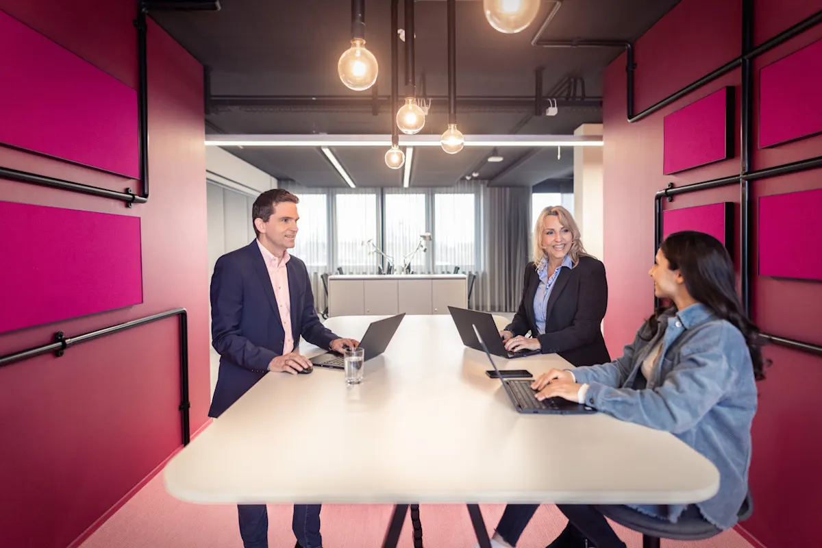 NEW AG - Verantwortung übernehmen Drei Personen sitzen in einem modernen Besprechungsraum an einem weißen Tisch mit Laptops und führen ein Gespräch. Der Raum ist in auffälligem Pink gestaltet mit auffälligen Leuchtmitteln und großen Fenstern im Hintergrund.