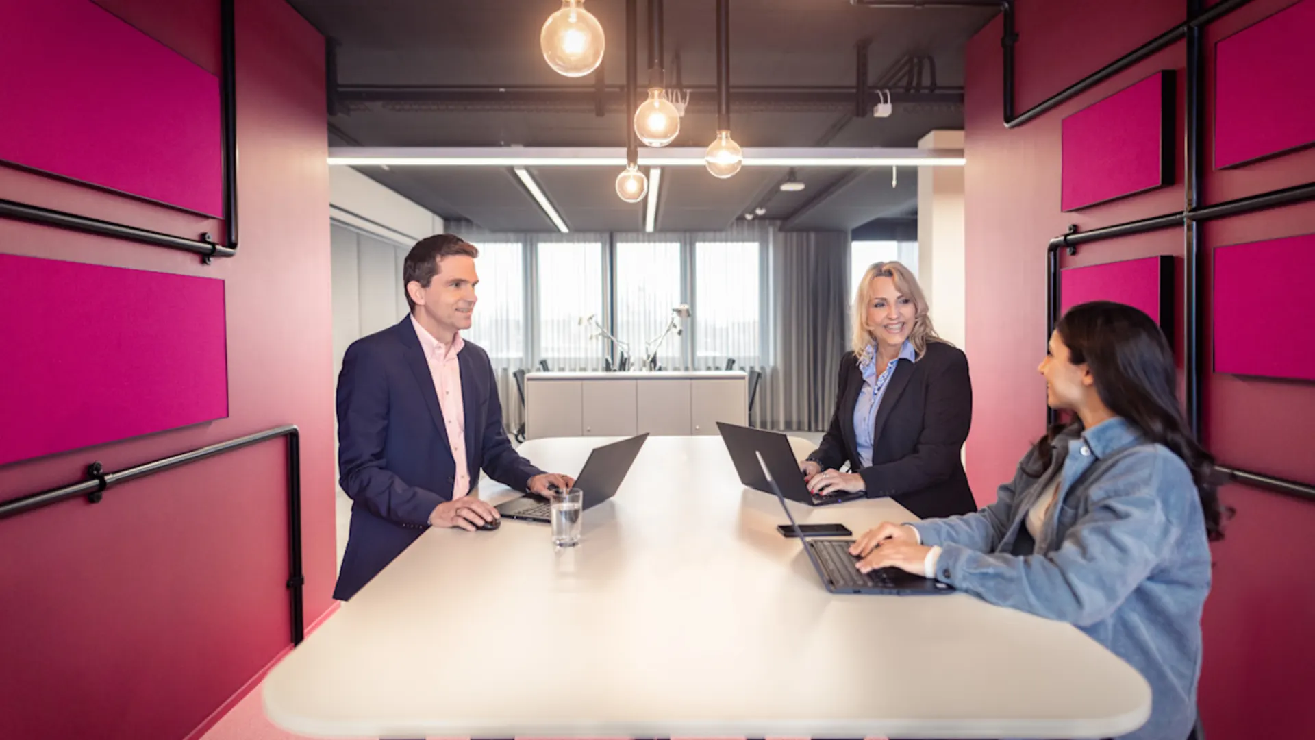 NEW AG - Verantwortung übernehmen Drei Personen sitzen in einem modernen Besprechungsraum an einem weißen Tisch mit Laptops und führen ein Gespräch. Der Raum ist in auffälligem Pink gestaltet mit auffälligen Leuchtmitteln und großen Fenstern im Hintergrund.