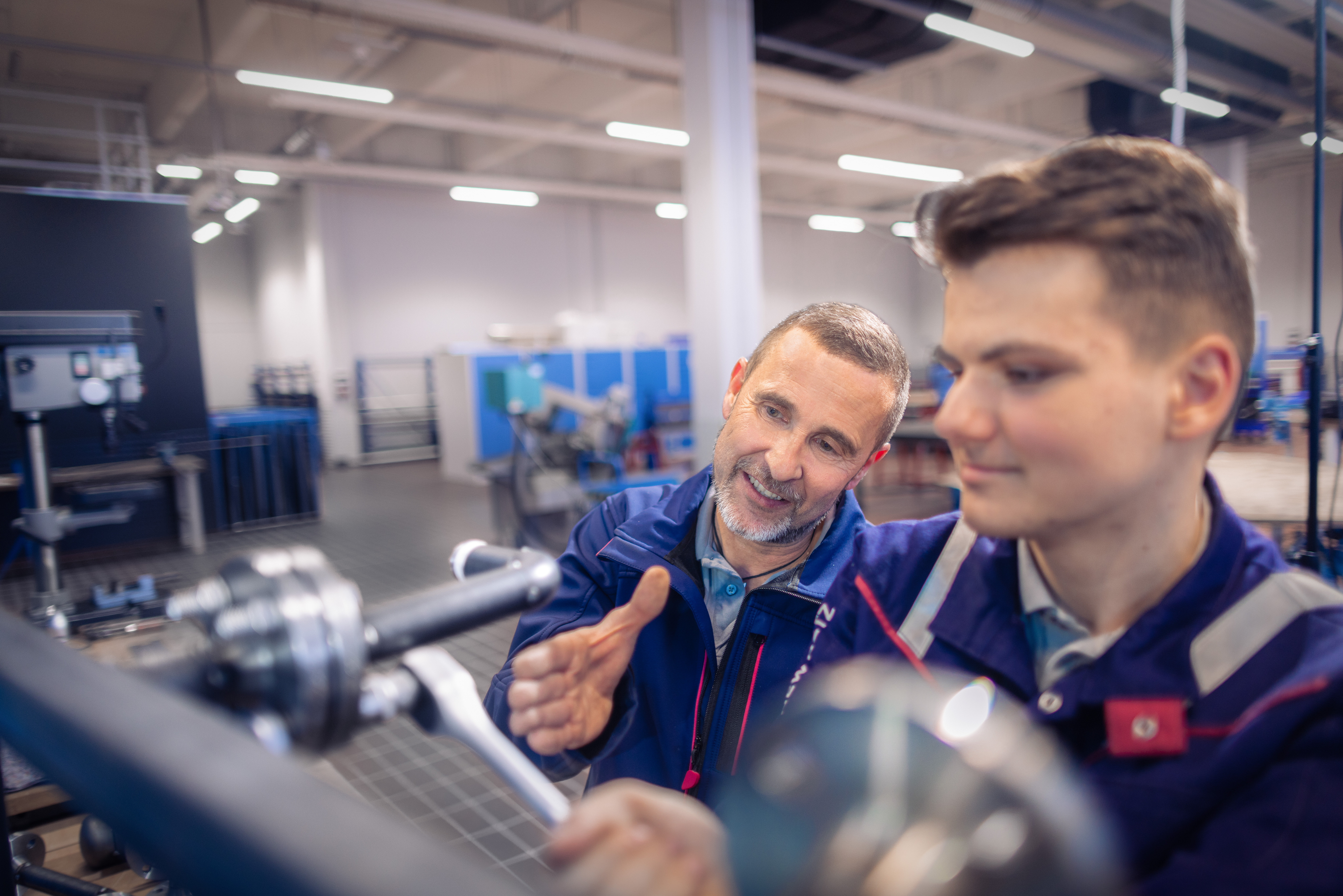 Ein Ausbilder in blauer Arbeitskleidung erklärt einem jungen Auszubildenden etwas an einer Maschine in einer modernen Werkstatt. Der ältere Mann deutet mit der Hand auf Details, während der junge Mann konzentriert auf ein technisches Gerät schaut. Im Hintergrund sind weitere Arbeitsbereiche und Geräte in der hell beleuchteten Industriehalle zu sehen.