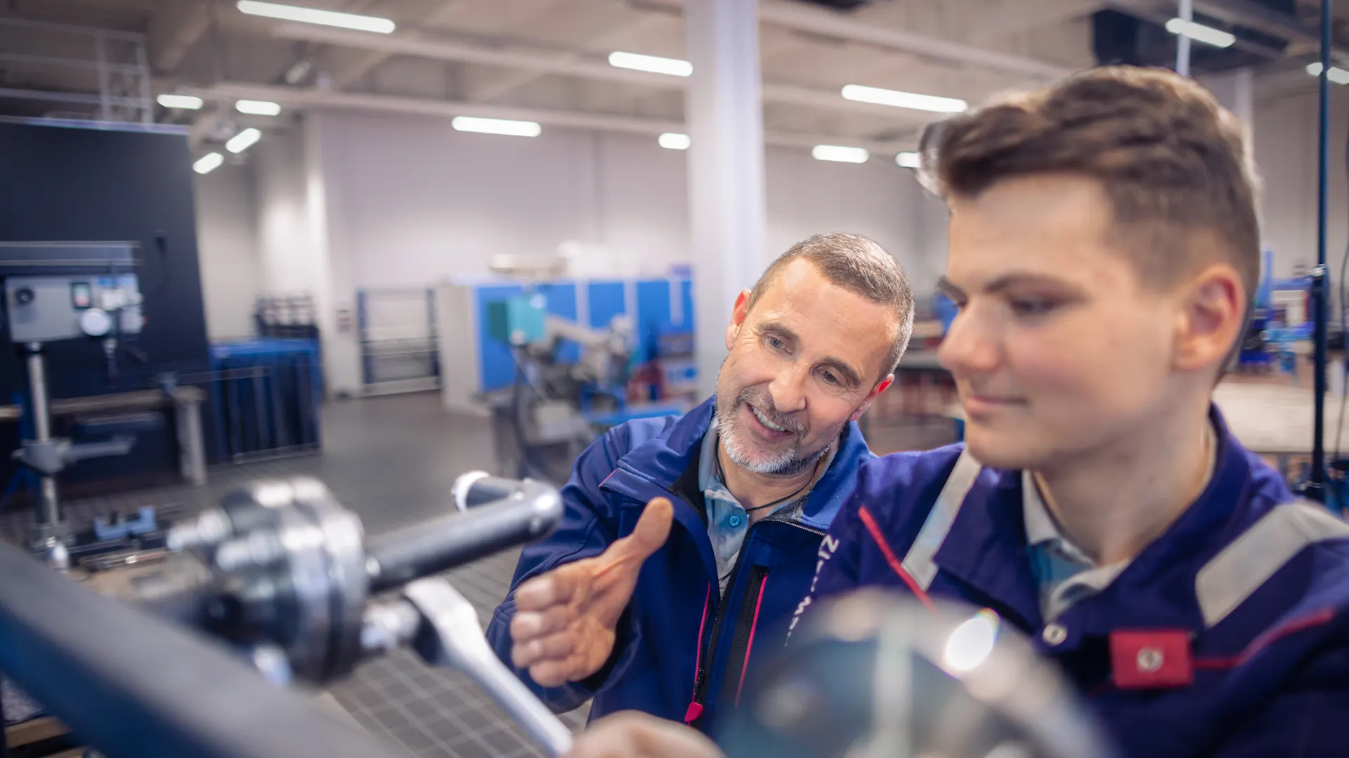 Technische Ausbildung Ein Ausbilder in blauer Arbeitskleidung erklärt einem jungen Auszubildenden etwas an einer Maschine in einer modernen Werkstatt. Der ältere Mann deutet mit der Hand auf Details, während der junge Mann konzentriert auf ein technisches Gerät schaut. Im Hintergrund sind weitere Arbeitsbereiche und Geräte in der hell beleuchteten Industriehalle zu sehen.