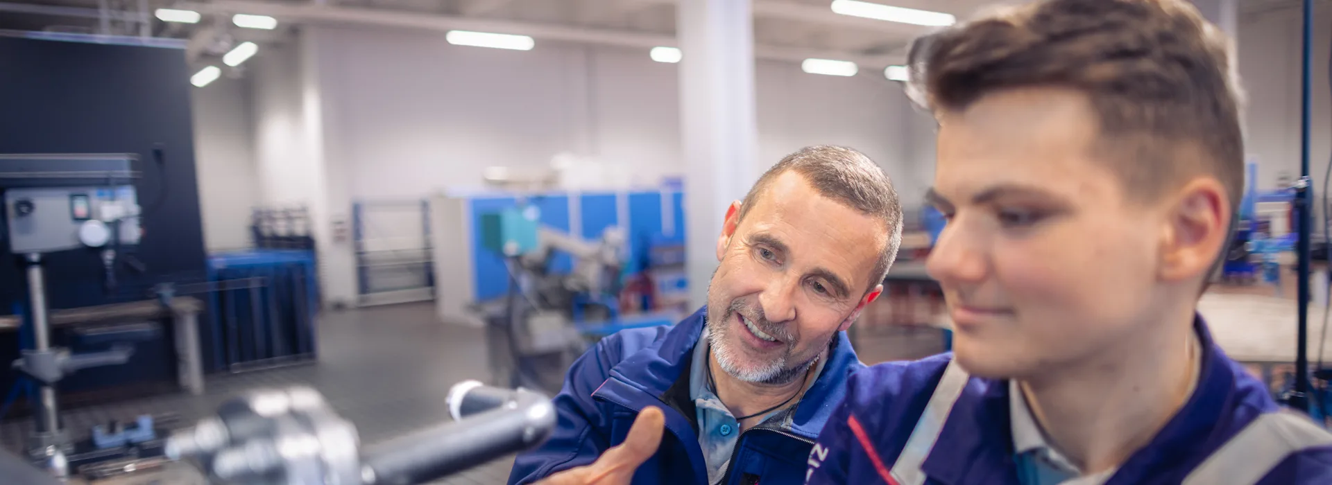 Technische Ausbildung Ein Ausbilder in blauer Arbeitskleidung erklärt einem jungen Auszubildenden etwas an einer Maschine in einer modernen Werkstatt. Der ältere Mann deutet mit der Hand auf Details, während der junge Mann konzentriert auf ein technisches Gerät schaut. Im Hintergrund sind weitere Arbeitsbereiche und Geräte in der hell beleuchteten Industriehalle zu sehen.