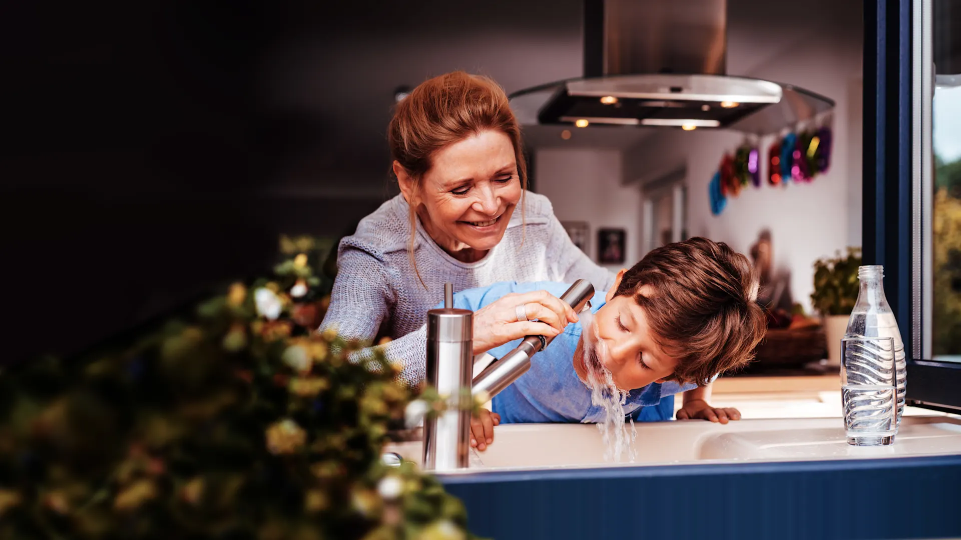 Kind trinkt aus dem Wasserhahn an einem Spülbecken Kind trinkt aus dem Wasserhahn an einem Spülbecken. Eine Frau steht dahinter