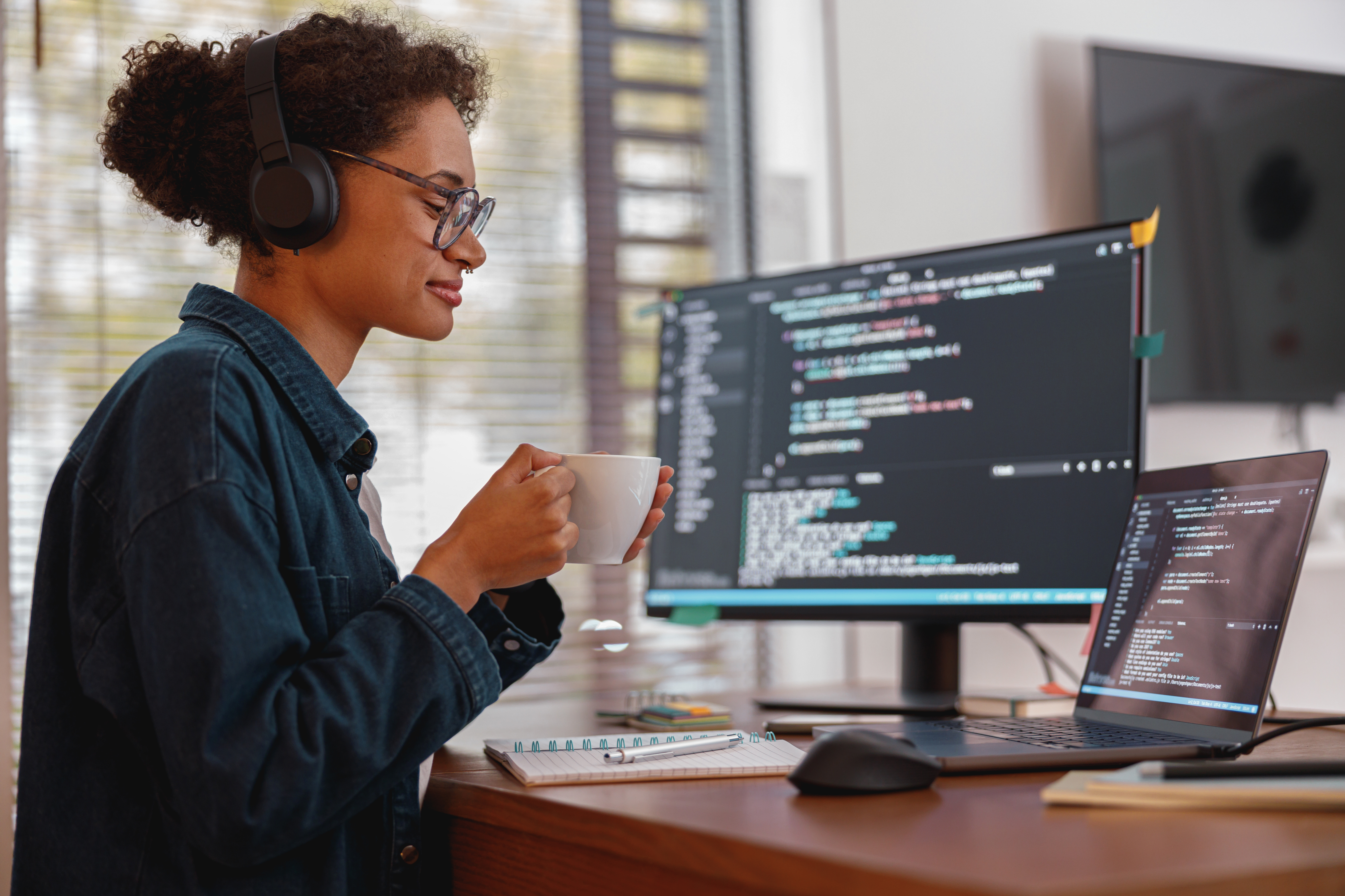 Person sitzt mit Kopfhörern vor einem Computerarbeitsplatz und hält eine Tasse in der Hand, während auf dem Bildschirm Programmcode Eine s zu sehen sind. Auf dem Schreibtisch liegen Notizen und ein Laptop. 