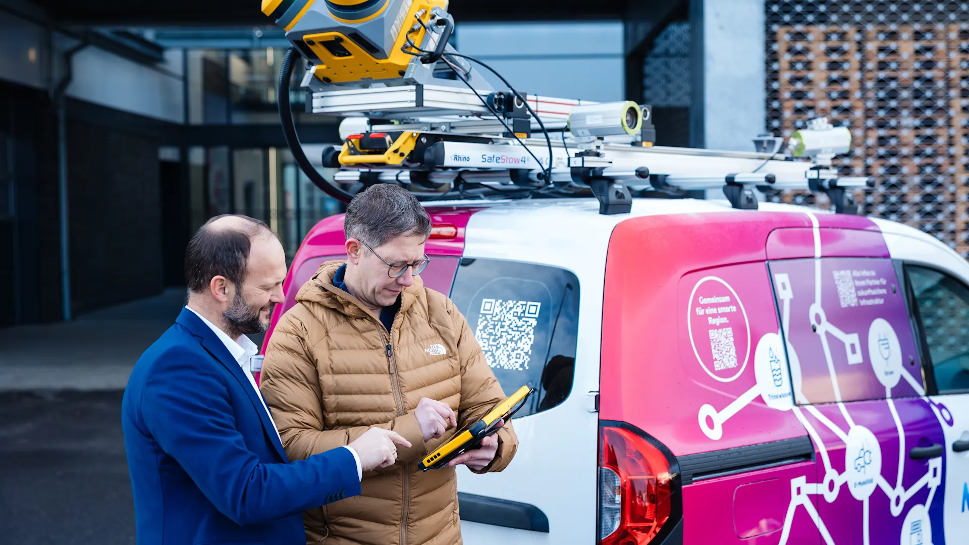 Mobile Mapping Fahrzeug Einsatzsituation im Außenbereich: Zwei Mitarbeitende stehen neben einem Servicefahrzeug mit auffälligem NEW‑Branding und technischer Ausstattung auf dem Dach. Dort ist ein spezialisiertes Mess‑ oder Erfassungssystem montiert. Eine Person hält ein mobiles Endgerät, während die andere unterstützend auf die Anzeige zeigt. Die Szene vermittelt moderne, digitale Arbeitsprozesse, mobile Datenerfassung und den Einsatz innovativer Technik im praktischen Einsatzfeld.