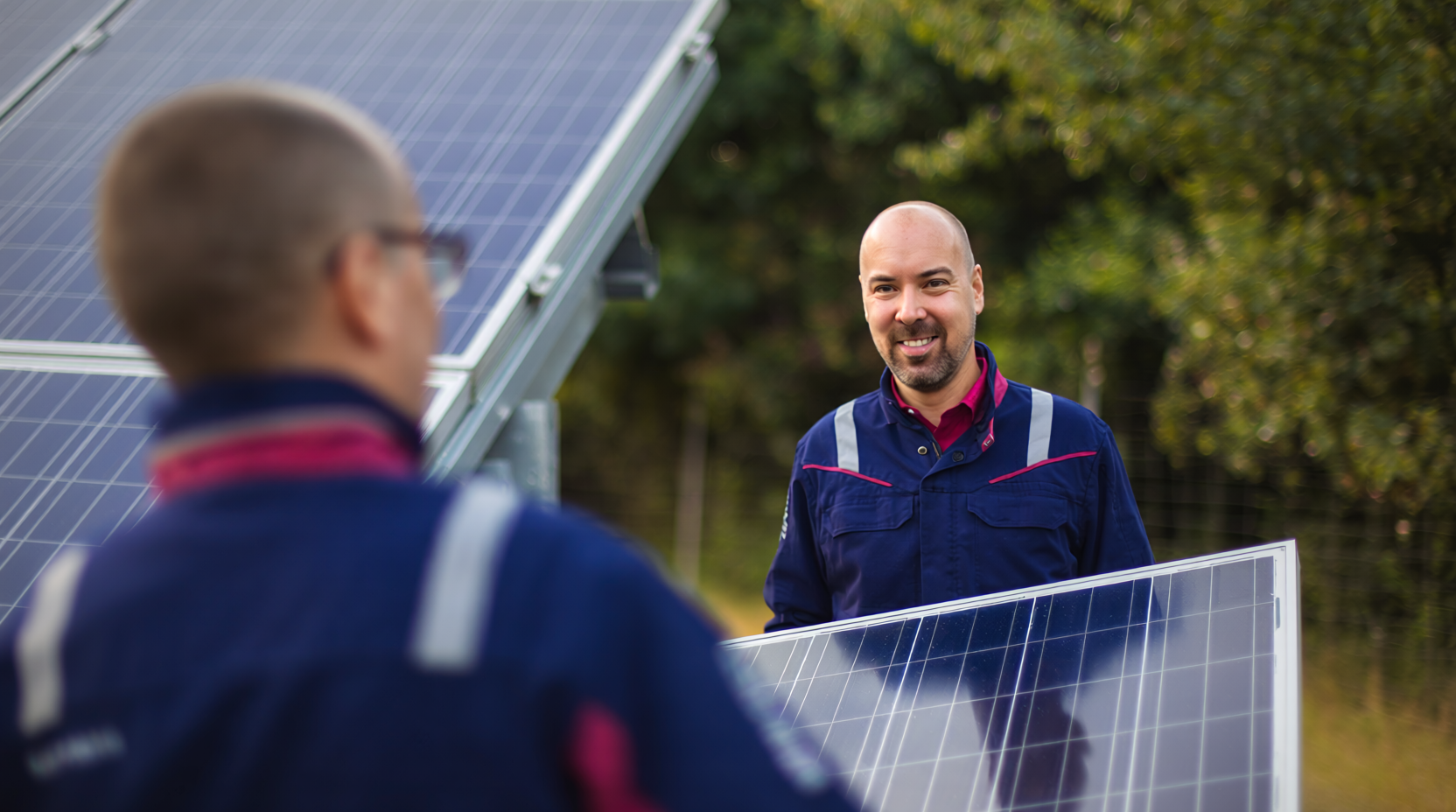 Zwei Techniker in blauer Arbeitskleidung mit reflektierenden Streifen arbeiten gemeinsam an der Installation von Solarmodulen im Freien. Im Hintergrund sind weitere Solarpaneele und grüne Bäume zu sehen.