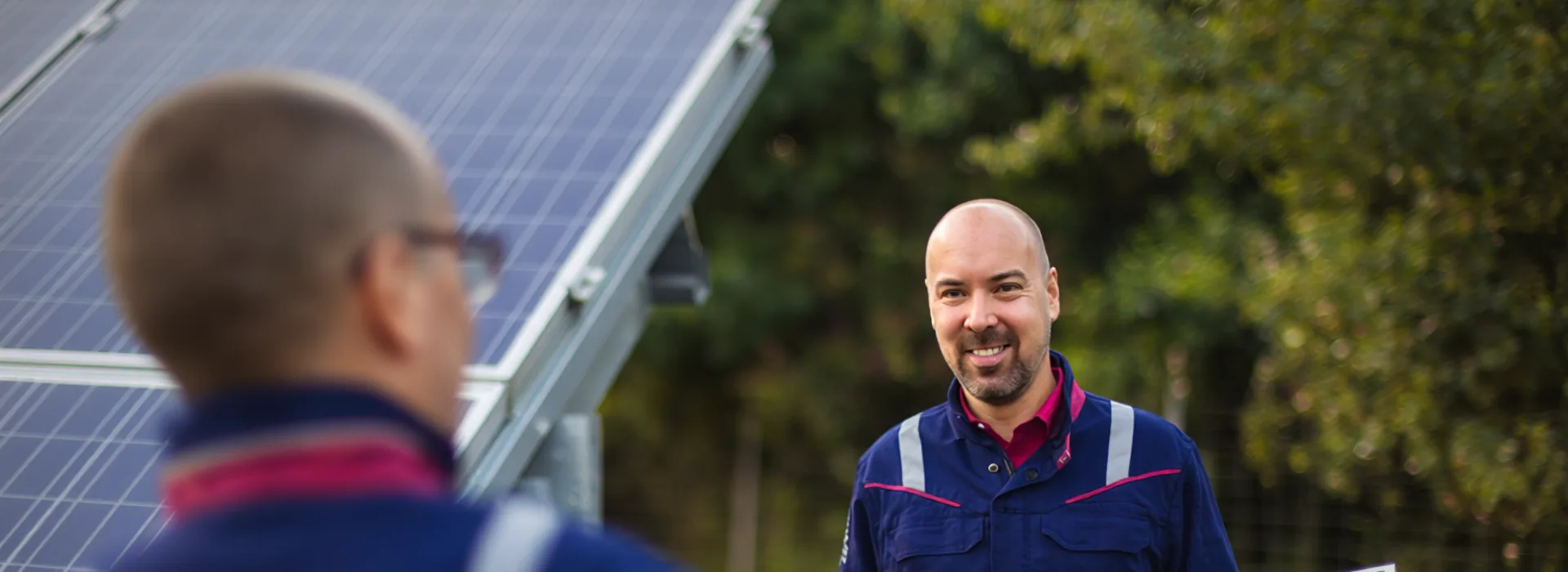 Berufseinssteiger - Zwei Mitarbeiter in einem Solarpark Zwei Techniker in blauer Arbeitskleidung mit reflektierenden Streifen arbeiten gemeinsam an der Installation von Solarmodulen im Freien. Im Hintergrund sind weitere Solarpaneele und grüne Bäume zu sehen.