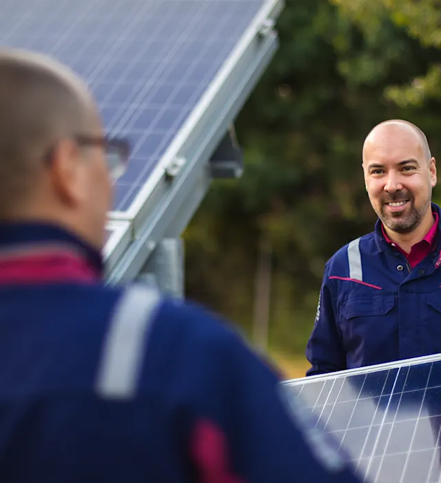 Berufseinssteiger - Zwei Mitarbeiter in einem Solarpark Zwei Techniker in blauer Arbeitskleidung mit reflektierenden Streifen arbeiten gemeinsam an der Installation von Solarmodulen im Freien. Im Hintergrund sind weitere Solarpaneele und grüne Bäume zu sehen.
