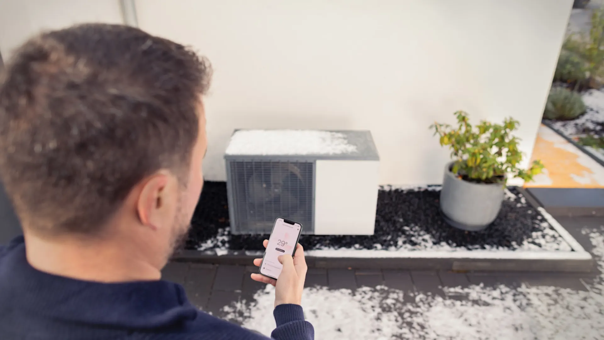 Der beste Weg zur nachhaltigen Wärmeversorgung. Ein Mann in dunklem Pullover bedient eine Wärmepumpe über eine Smartphone-App. Auf dem Handy-Display ist eine Temperaturanzeige sichtbar. Die moderne kleine Wärmepumpe steht auf einem Kiesbett neben einem grauen Blumentopf mit grüner Pflanze.