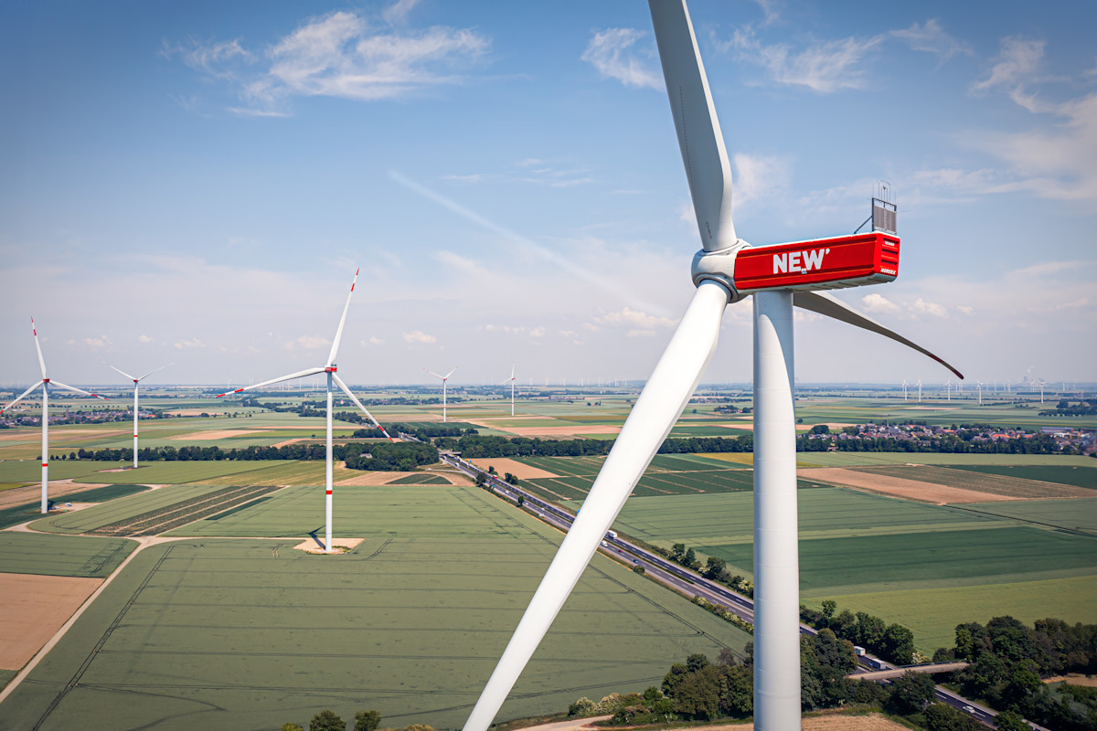 Luftaufnahme von modernen Windkraftanlagen in ländlicher Landschaft. Im Vordergrund steht eine große Windturbine mit dem roten NEW-Logo am Turm, weitere Windräder sind in der Ferne sichtbar.