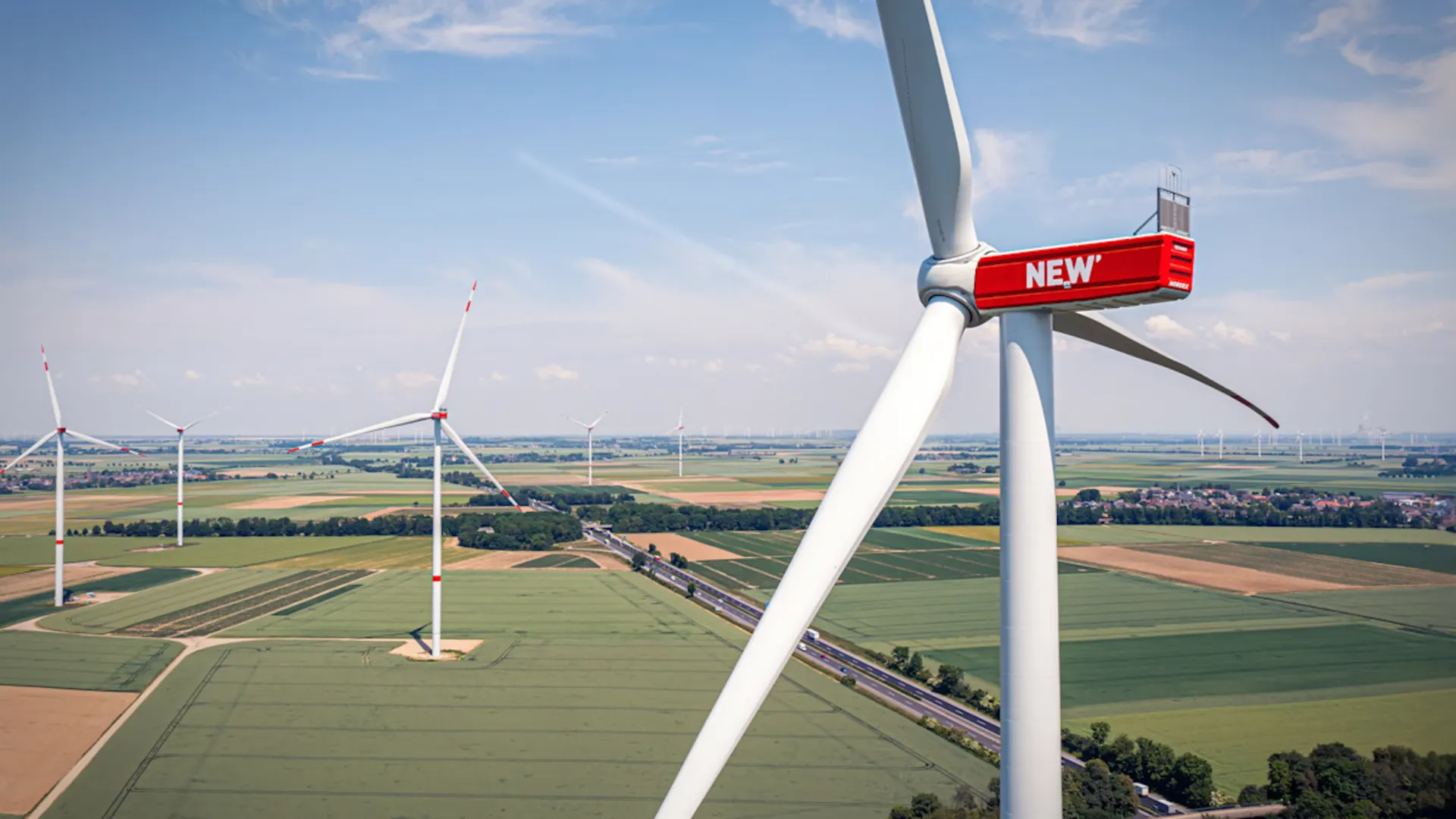 Energie von morgen, regenerativ gedacht. Luftaufnahme von modernen Windkraftanlagen in ländlicher Landschaft. Im Vordergrund steht eine große Windturbine mit dem roten NEW-Logo am Turm, weitere Windräder sind in der Ferne sichtbar.