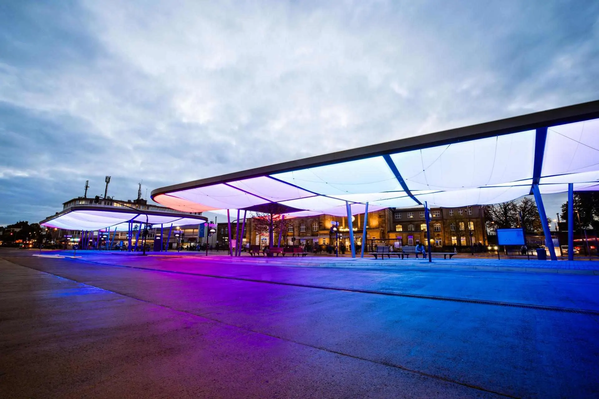 Das Bild zeigt den Zentralen Omnibusbahnhof (ZOB) in Mönchengladbach in der Dämmerung. Die moderne Dachkonstruktion ist farbig beleuchtet und prägt das Stadtbild. Im Hintergrund befindet sich der Hauptbahnhof