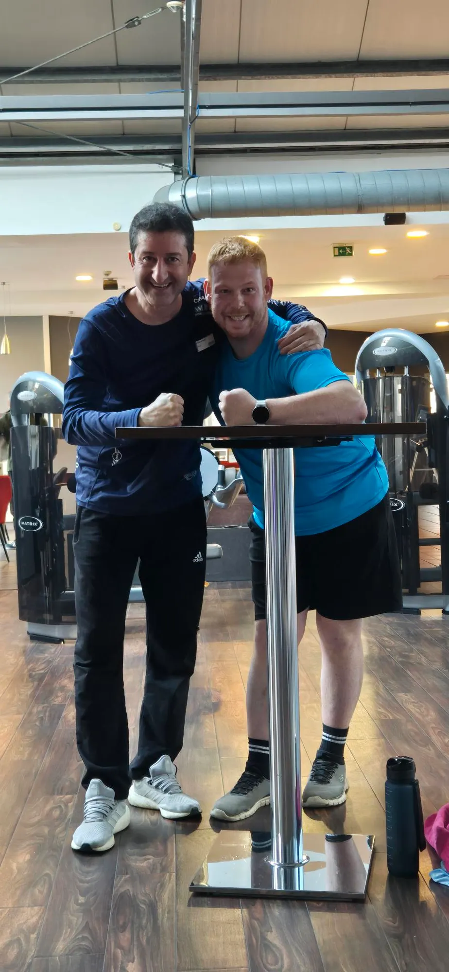 Martin Giesers-Aviles in blauem Sportshirt steht Arm in Arm mit seinem Trainer Medi in dunklem Shirt an einem Trainingsgerät im Fitnessstudio und beide lächeln in die Kamera.