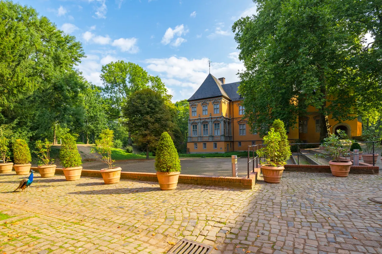 Gepflasterter Innenhof eines historischen orangefarbenen Gebäudes, umgeben von Laubbäumen. Im Vordergrund ist ein Pfau zu erkennen. Der Himmel ist blau mit einzelnen Wolken.  