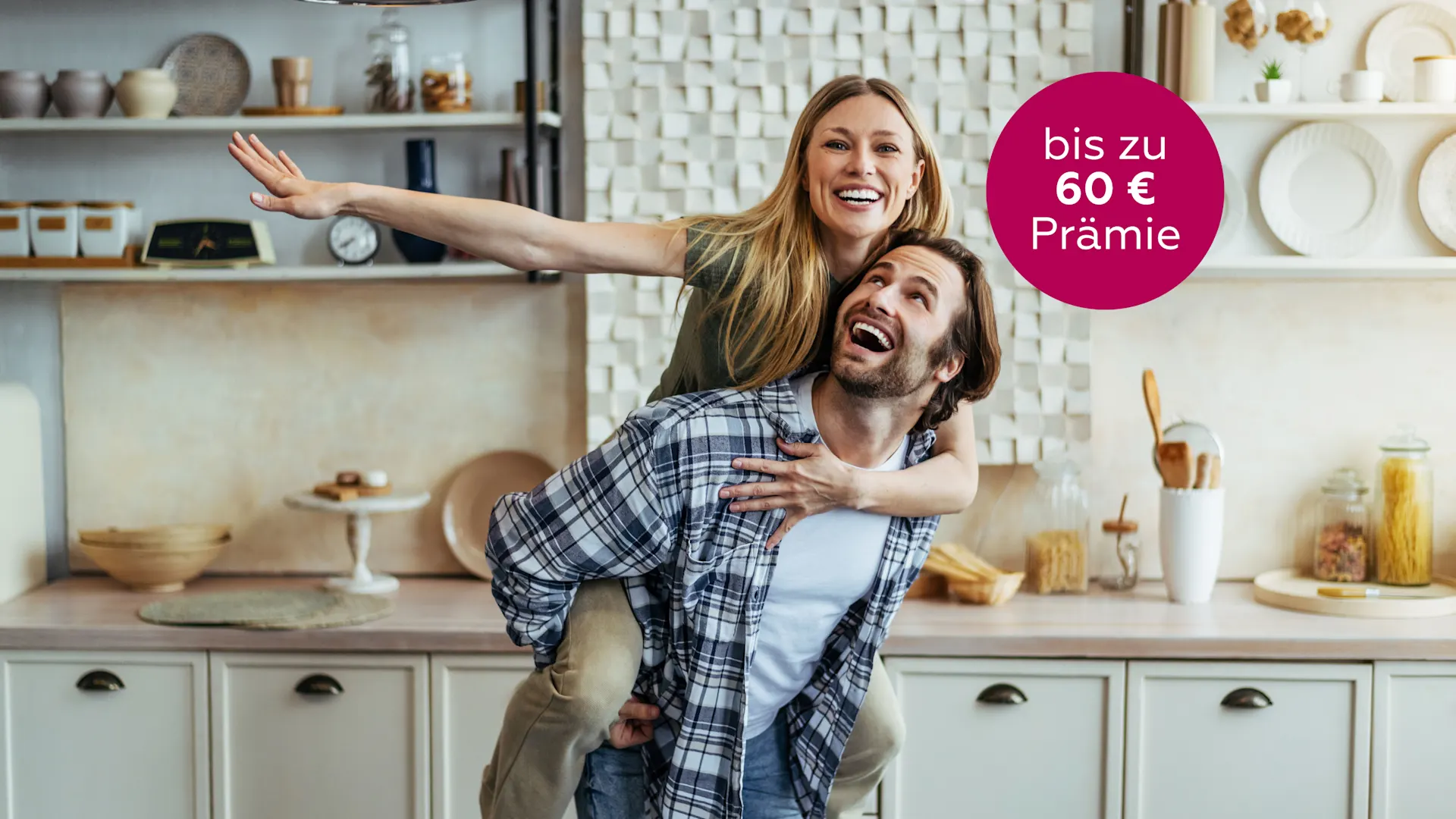 Jetzt NEW Energie empfehlen und 50 € Prämie sichern Mann trägt eine lachende Frau Huckepack auf dem Rücken und steht in einer Küche.