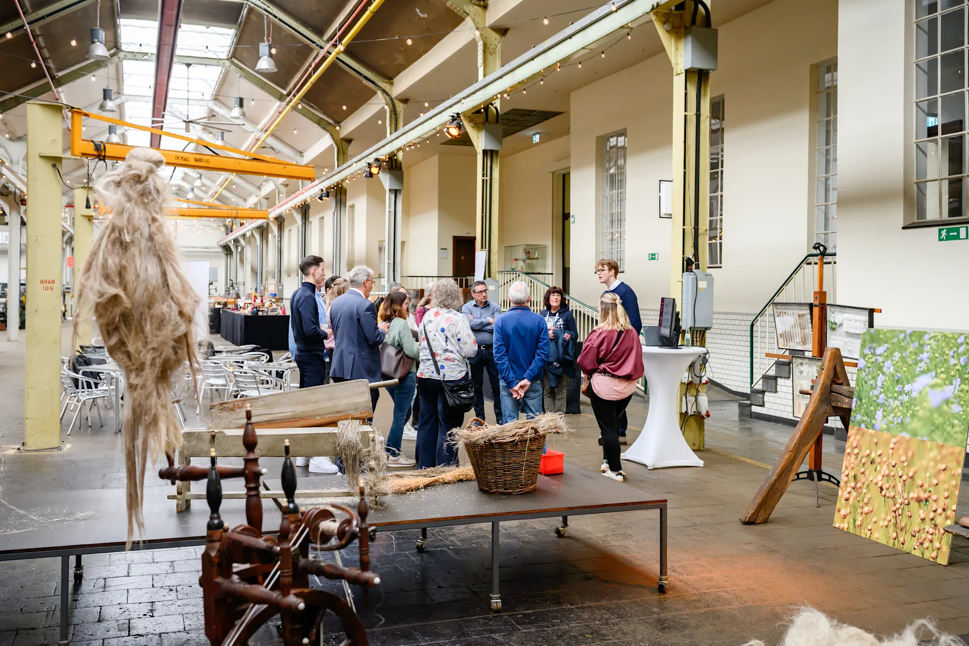 Menschen stehen in einer lichtdurchfluteten Industriehalle zusammen und unterhalten sich. Im Vordergrund sind ein Spinnrad und Naturfasern auf einem Tisch zu sehen, im Hintergrund Stehtische und ein Gemälde mit Blumenmotiv.