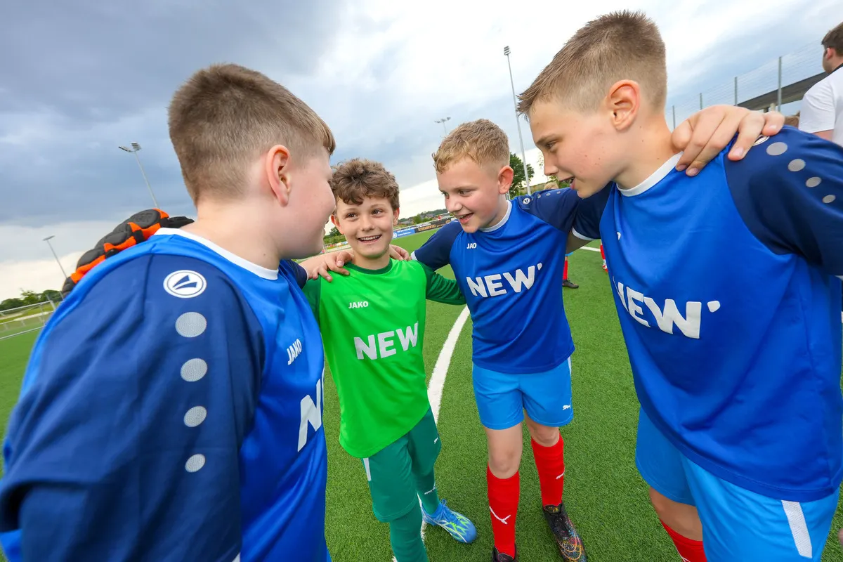 Trikotsponsoring: Teamgeist sichtbar machen Junge Fußballspieler in blauen NEW-Trikots stehen Arm in Arm auf einem Fußballplatz. Ein Junge trägt ein grünes Torwarttrikot, alle anderen sind in blau gekleidet. Die Kinder lächeln sich an und strahlen Teamgeist und Zusammenhalt aus.