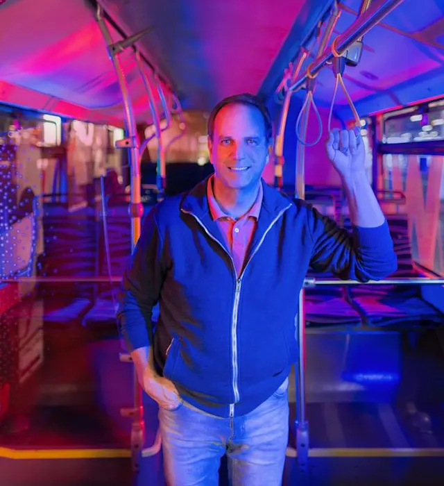 NEW MöBus Ein Mann steht im Bus, lächelt in die Kamera und hält sich am Haltegriff fest.