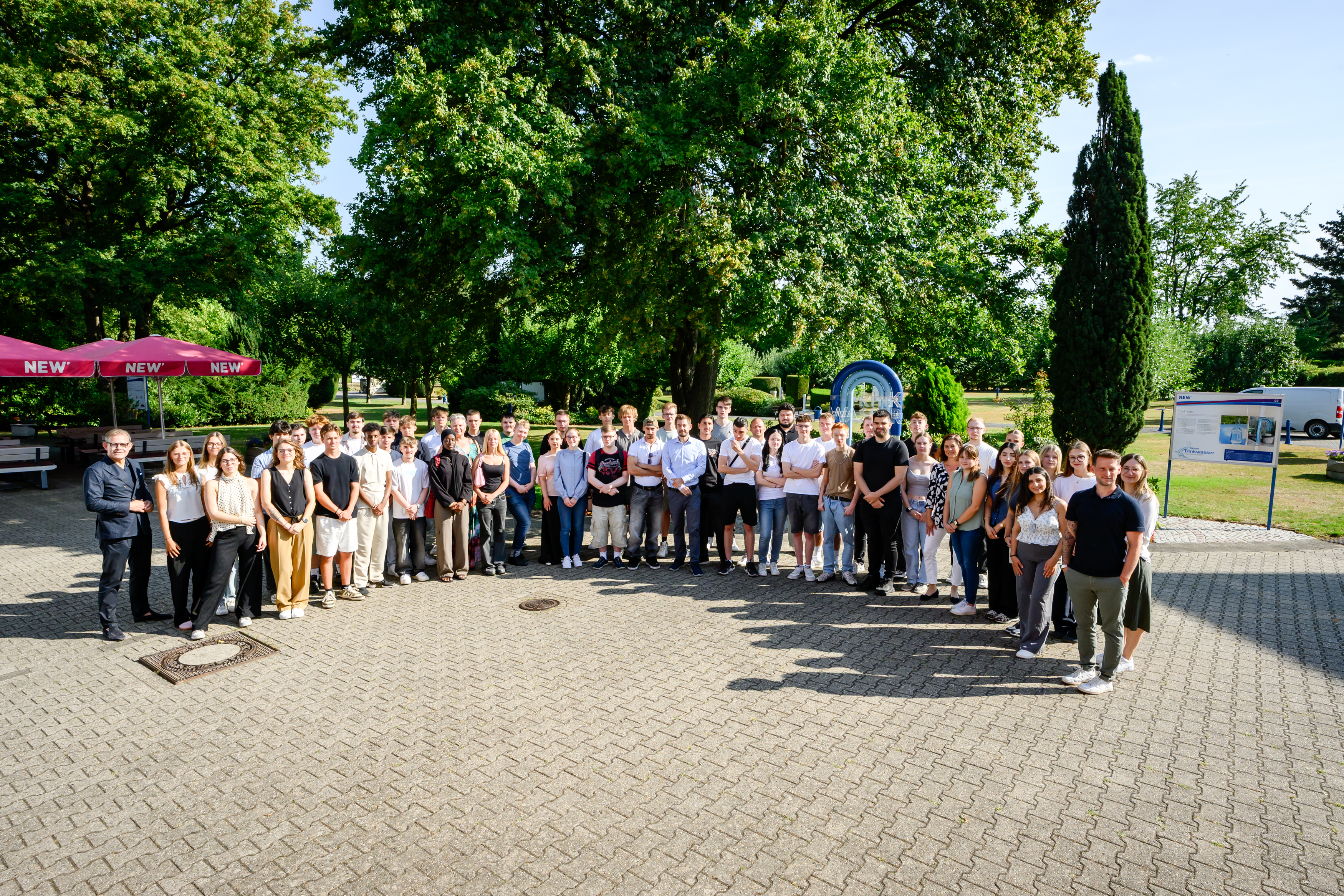 Gruppenfoto einer großen Anzahl neuer Auszubildender sowie beteiligter Mitarbeitender der NEW auf einem befestigten Platz im Außenbereich. Die Teilnehmenden stehen in mehreren Reihen vor einer grünen Park- und Gartenkulisse mit großen Bäumen. Links ist ein roter Sonnenschirm mit NEW-Branding zu sehen. Das Foto zeigt das gemeinsame Zusammenkommen im Rahmen einer Willkommens- oder Einführungsveranstaltung für den Ausbildungsstart bei der NEW. Die Aufnahme vermittelt den Gemeinschaftsgedanken, den persönlichen Austausch und den Start in die berufliche Zukunft innerhalb der NEW-Gruppe.