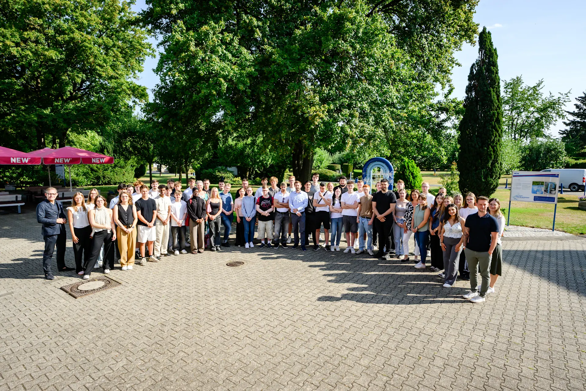NEW Azubi Willkommen-tung 03.08.2026 Gruppenfoto einer großen Anzahl neuer Auszubildender sowie beteiligter Mitarbeitender der NEW auf einem befestigten Platz im Außenbereich. Die Teilnehmenden stehen in mehreren Reihen vor einer grünen Park- und Gartenkulisse mit großen Bäumen. Links ist ein roter Sonnenschirm mit NEW-Branding zu sehen. Das Foto zeigt das gemeinsame Zusammenkommen im Rahmen einer Willkommens- oder Einführungsveranstaltung für den Ausbildungsstart bei der NEW. Die Aufnahme vermittelt den Gemeinschaftsgedanken, den persönlichen Austausch und den Start in die berufliche Zukunft innerhalb der NEW-Gruppe.