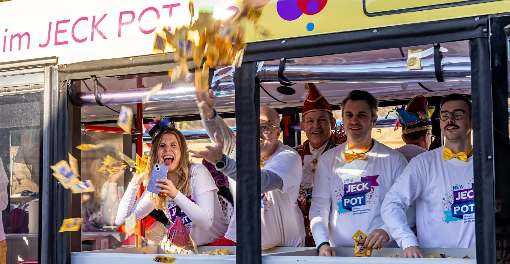 Eine Frau wirft fröhlich aus einem Karnevalswagen Tüten mit Gummibärchen in die Luft. Mehrere lächelnde Personen in weißen Shirts stehen im Wagen und verteilen Süßigkeiten.  Credit: NEW AG/Christopher Lambers