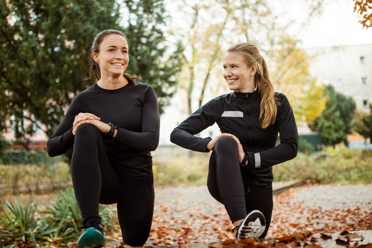 Zwei Frauen in sportlicher Kleidung stretchen sich in einem Park.