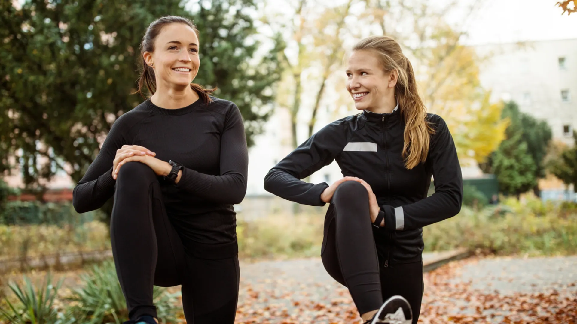 Bewegung in die Mittagspause bringen: So geht’s Zwei Frauen in sportlicher Kleidung stretchen sich in einem Park.